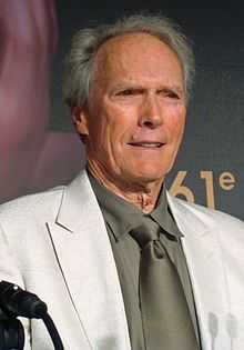 An older man is at the center of the image smiling and looking off to the right of the image. He is wearing a white jacket, and a tan shirt and tie. The number 61 can be seen behind him on a background wall.