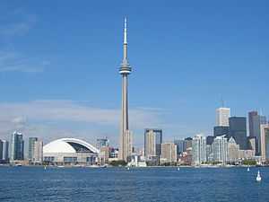 A spear-like tower is between a white-domed structure and small buildings on the left, and increasingly taller buildings to the right.  In the foreground is a lake, with a few visible buoys, and the background is a deep blue sky with a few clouds near the horizon.