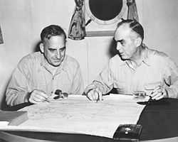 Two men sit at a small table. A map is laid out on the table. Its four corners are held down by a small propeller, a glass, and a book.