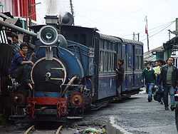 A train, hauled by a steam locomotive, running beside a road between two rows of buildings with a few people walking on the road.