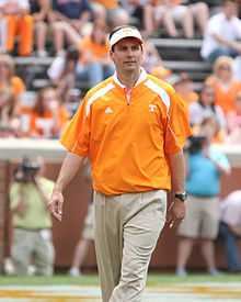 Coach Dooley smiles at practice in an orange shirt and hat on the sideline of an American football field. At rear are several persons with cameras and fans in the stadium seats.