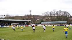 Two teams competing in a football match, one weraing white shirts and black shorts and the other yellow shirts and blue shorts.  A stand full of spectators and another low brick building are visible outside the playing area.