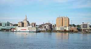 A row of tall buildings in the middle, with a river on the bottom and a blue cloudy sky on top