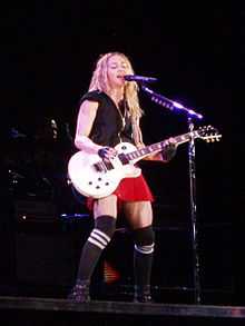 A faraway image of a blond woman, wearing a red shorts, black T-shirt and playing a white electric guitar. Her face is away from the image. Beside her, a band stands with the backdrops displaying white stars on black.