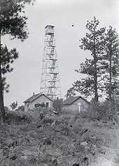 A four-legged tower with a small  at the top, next to two one-story buildings. The tower is four stories tall. Trees are at either side, and in the foreground there are rocks, some vegetation, and a rough trail.