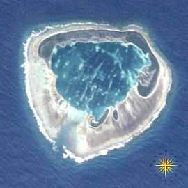 A satellite photograph of an atoll consisting of four islets with an interior lagoon. The largest island has a "C" shape and is dominated by vegetation. The other three are smaller and have sparse vegetation. The islets are surrounded by dead coral and the ocean. In the lower right corner of the photo, a compass indicates the orientation of the island.