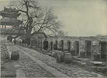 A black-and-white picture of a stone-paved alley going from bottom right to top left leading to a three-roofed gate and bordered on the right by a line up of small roofed cubicles open on one side.