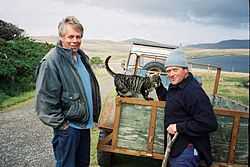 Photograph of two men and a cat standing next to a truck on the side of a road