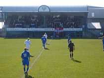 A football match with players and a referee on the pitch. A small spectator stand full of people is in the background.