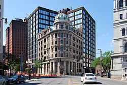 An older, ornate stone and brick building with a small domed green top in a city. It is seen from a street that goes straight toward it, looking directly at its curved corner. There are larger, more modern buildings behind it.