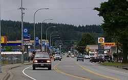 Photo of a three-lane main street in a small town. Visible is a traffic light, sparse traffic, and businesses such as a car lot and gas station.