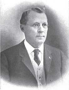 A man with wavy, graying hair wearing glasses, a white shirt, light vest, and a dark tie and jacket