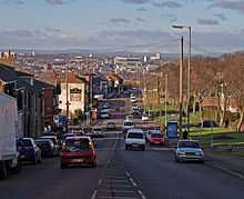 Ina suburban area, a busy main road wit vehicles travelling in both directions recedes into the distance. On the left is a row of red-bricked two-storey houses. On the right, an open parkland fringed with mature trees is visible. In the distance is a sunlit landscape of urban and industrial buildings leading to distant, low hills.