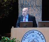 A man in his fifties standing behind a desk with computers on it. On the desk is a sign reading "Kungl. Vetensk. Akad. Sigil".