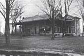 A house of wooden construction with a wrap-around front porch and trees in the front yard