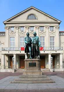 Photo of a statue of two men in front of a large stone building with a columnade at the entrance, many rectangular windows and carved decorations at roof level