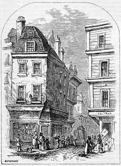 People congregate at the entrance to a narrow street, overlooked by two four-storey buildings.  Each floor of the right-most building projects further over the street than the floor below.  At the corner of each building, shops advertise their wares.  A cart is visible down the street, and one man appears to be carrying a large leg of meat.