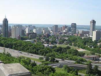 A tree-lined highway is in the foreground, angled diagonally from bottom right to middle left of the image. Buildings are in the centre, and the background is a sky meeting rolling hills in the distance.