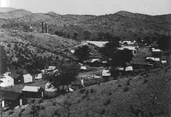 A street of about 30 one-story buildings stretches along the floor of a dry, narrow valley. A few trees grow near the houses, but only low, widely separated desert shrubs grow on the hills above the valley. Two smokestacks are visible just over the nearest hill.