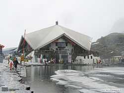 A stone building is surrounded by partially frozen ponds. Pilgrims can be seen on the paths