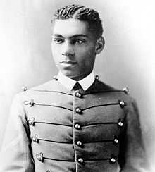 Cadet Henry O. Flipper in his West Point cadet uniform. It has three large round brass buttons left, middle and right showing five rows. The buttons are interconnected left to right and vice-versa by decorative thread. He is wearing a starched white collar and no tie. He is a lighter colored African-American with plated corn rows of neatly done hair. He is facing the camera and looking to the left of the viewer.