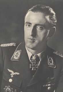 The head and shoulders of a man, shown in semi-profile. He wears a military uniform with various military decorations and an Iron Cross at the front of his shirt collar. His hair is dark and short and combed back, his nose is long and bent and his mouth is thin; he is looking into the camera