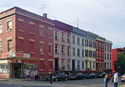 A row of four three-story flat-roofed brick buildings in various colors seen from across a corner. There are people standing on the other corners. The building nearest the camera, at the left, has a a sign at street level saying "Grand Deli" and another, smaller one saying "ATM".