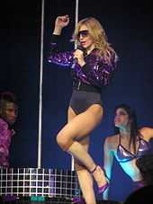 Image of a blond women. She's wearing a purple sparkly jacket and a purple leotard underneath. She's also wearing sunglasses and holds a microphone to her mouth. To her right there's a brunette female wearing a purple bra and to her left an African American male