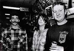 Three men standing side-by-side. A sign above the man on the left reads "Jazz!"