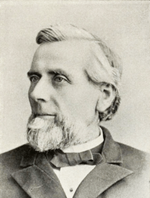 Head and shoulders photograph of a man in an overcoat.  He has a salt and pepper goatee and has his hair combed to the side.  He wears a calm expression and is looking slightly to the right.