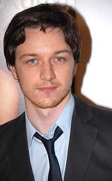 An up-close picture of a blue-eyed dark-haired Caucasian man in his early 30s. The man is wearing a dark blue shirt and looking into the camera lens. People, trees and a tall building can be seen in the background.