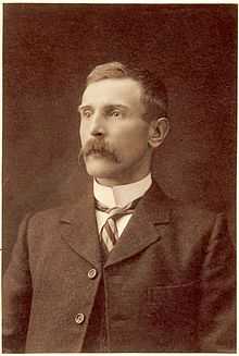 Head and shoulders portrait of a man with a moustache in a suit and tie with a high collar.