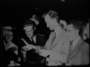 Waist high portrait of male in his forties, quite poor quality, taken before sunrise, wearing a light colored suit. Man in uniform to his right, onlooker at right.