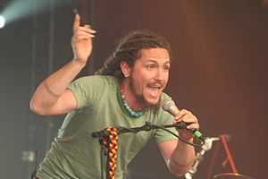 A thirty-one year-old man is shown in upper-body shot and slightly on his right profile. He is leaning forwards with his left hand grasping a microphone as he sings into it. His right arm is raised above his head and shows his long fingernails. Both fore-arms show a similar tattoo of three encircling lines. He wears an ear-ring in his right lobe, a necklace of red and blue-green beads and a green T-shirt. He is bearded and the start of his dreadlocks is visible.