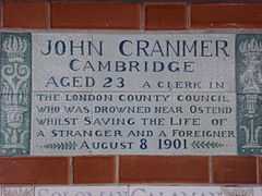 A tablet formed of six standard sized tiles, bordered by green flowers in the style of the Arts and Crafts movement. The tablet reads "John Cranmer Cambridge aged 23, a clerk in the London County Council who was drowned near Ostend whilst saving the life of a stranger and foreigner, August 8, 1901".