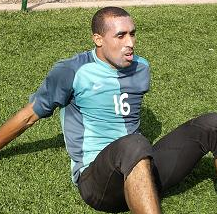 A football player in his number 16 goalkeeper's kit, sitting on the pitch with knees up and arms stretched back