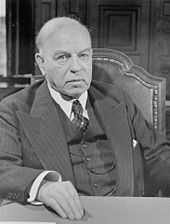 An older white man in a pinstriped three piece suit sits facing the camera, his right hand resting in front of him on a table.