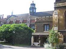  A long, low building of brick and stonework, with arched windows. It is partially obscured by trees and shrubs. The slate roof features a six-sided tower of stone and class.