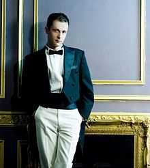 A dark haired man in a tailcoat and a kummerbund, dark bow tie, both hands in the pockets of light-coloured trousers.