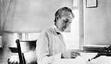 Woman sitting at desk writing, with short hair, long-sleeved white blouse and vest