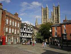 Aviw of Castle square - which is not a square at all.  In the foreground is the cobbled surface of the open space which is surrounded by historic buildings.  From left to right, the regency assembly rooms, a georgian 3 story house, then Leigh-Pemberton house: a half-timbered building with the upper story jettied out.  Then a medieval church and the entrance to the Cathedral cclose.  In the left distance the towers of the cathedral rise into a blue sky.