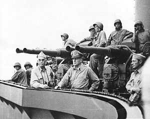 Sailors wearing steel helmets stand by an anti-aircraft gun on a quadruple mount. Two officers lean on the railing, staring off into the distance.