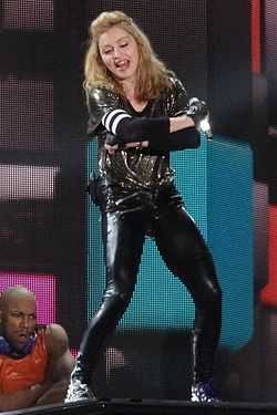 A blond woman in tight black spandex and a loose, glittering shirt, with gloves on her hand and elbow, poses ontage.