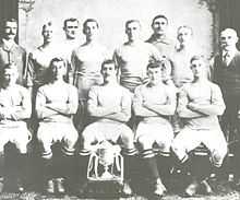 A group of thirteen men, eleven in association football attire typical of the early twentieth century and two in suits. A trophy sits in front of them