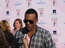 A black man, wearing sunglasses and a chequered shirt, speaks into a microphone whilst standing in front of a backdrop covered in sponsors' logos.