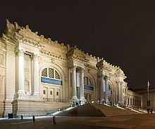 A very ornate multi-story stone façade rises over steps and a plaza at night.