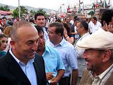 Two older Turkish men stand facing each other, one bald, the other wearing a white cap, while a large crowd mingles behind them along a waterfront.