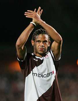 African man in a diagonally striped brown-and-white jersey with a sponsor logo across the chest. His hair is cornrowed, and his arms are crossed above his head.