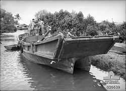 Four shirtless men in a large barge moored at a river bank. The barge has a ramp at the front of a catamaran hull.