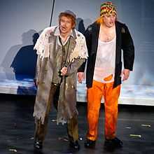 Two men in torn clothes stand on a stage: the man on the left is wearing a long coat and has a hat on, the man on the right wears a chequered hat, a string vest, jacket and orange trousers